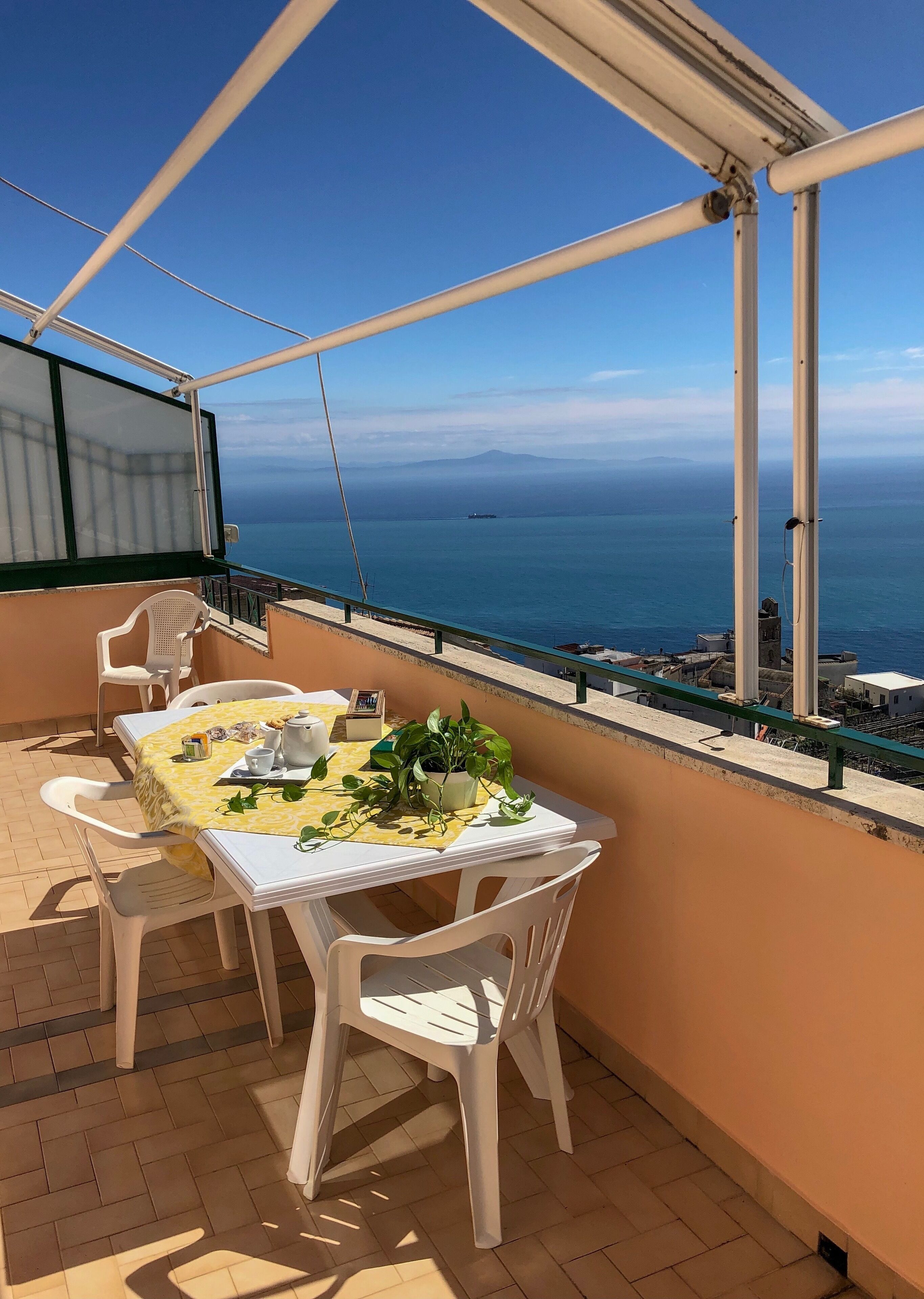 A balcony with a table set for two, white plastic chairs, a yellow tablecloth, and a potted plant.