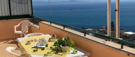 A balcony with a table set for two, white plastic chairs, a yellow tablecloth, and a potted plant.