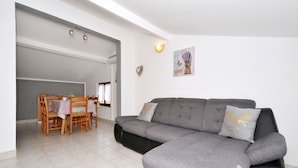 A modern living room with a grey sectional sofa, a dining table with chairs, and a kitchen area visible through an open door.