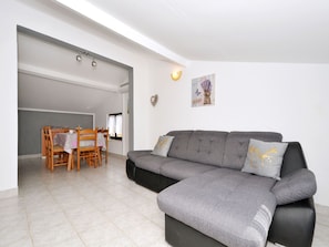 Una sala moderna con un sofá seccional gris, una mesa de comedor con sillas y una cocina visible a través de una puerta abierta.