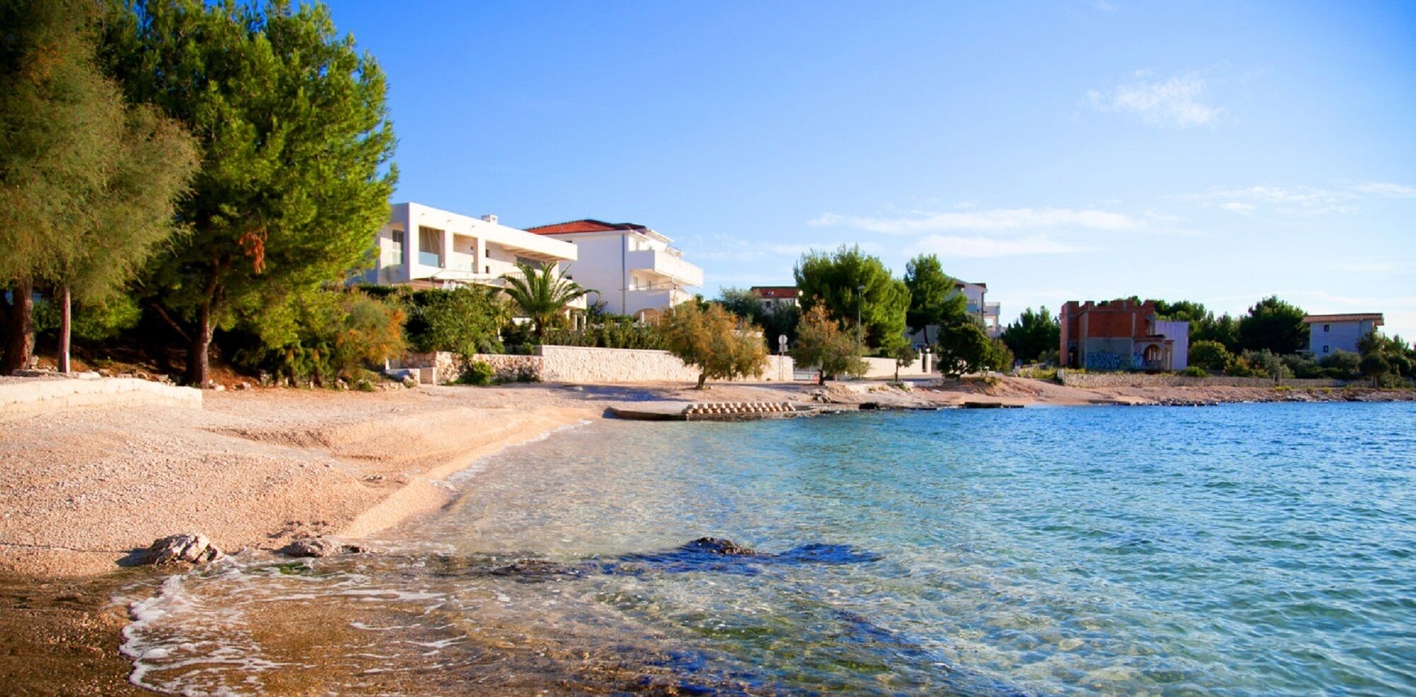 Eine Küstenlandschaft mit Sandstrand, klarem blauen Wasser und Gebäuden mit Balkonen mit Meerblick.