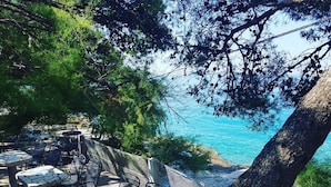 Outdoor seating with tables and chairs, overlooking a coastal area with trees and a view of the sea.