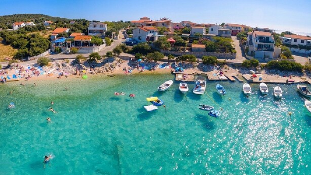A coastal village with clear turquoise waters, boats docked along the shore, and buildings with red-tiled roofs.