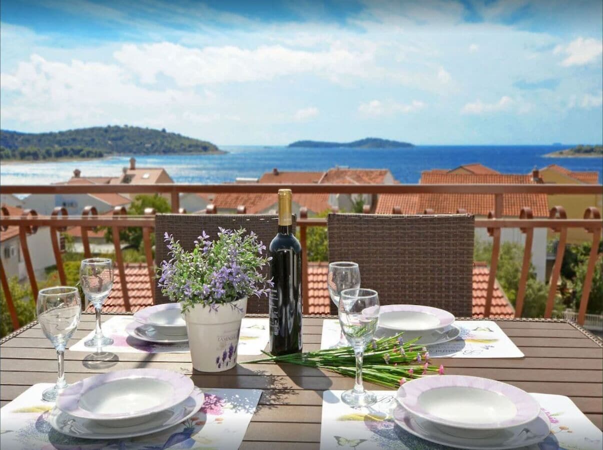 A table set for two with plates, glasses, a bottle of wine, and a potted plant on a balcony overlooking a scenic view.