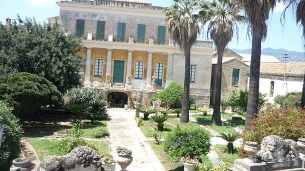 A building with a symmetrical facade, a central entrance with a portico, and a garden with palm trees and shrubs.