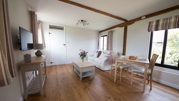 A cozy living room with a wooden floor, a white sofa, a dining table with chairs, a TV mounted on the wall, and a window with curtains. - Tastefully Renovated Countryside Cottage with Swimming Pool near Tenterden (High Halden)