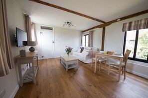 A cozy living room with a wooden floor, a white sofa, a dining table with chairs, a TV mounted on the wall, and a window with curtains. - Tastefully Renovated Countryside Cottage with Swimming Pool near Tenterden (High Halden)