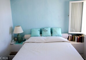 A bedroom with a bed, pillows, a nightstand, a lamp, and a window with books on the sill.