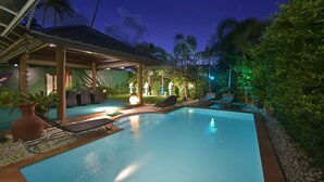 A pool area with lounge chairs and a covered seating area, surrounded by greenery and palm trees.