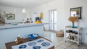 A kitchen with a white countertop, a dining table with a blue tablecloth, and a white cabinet with bottles. - Villa Paros - Archipelagos Villas with Pool close to the sea (Naousa Paros)