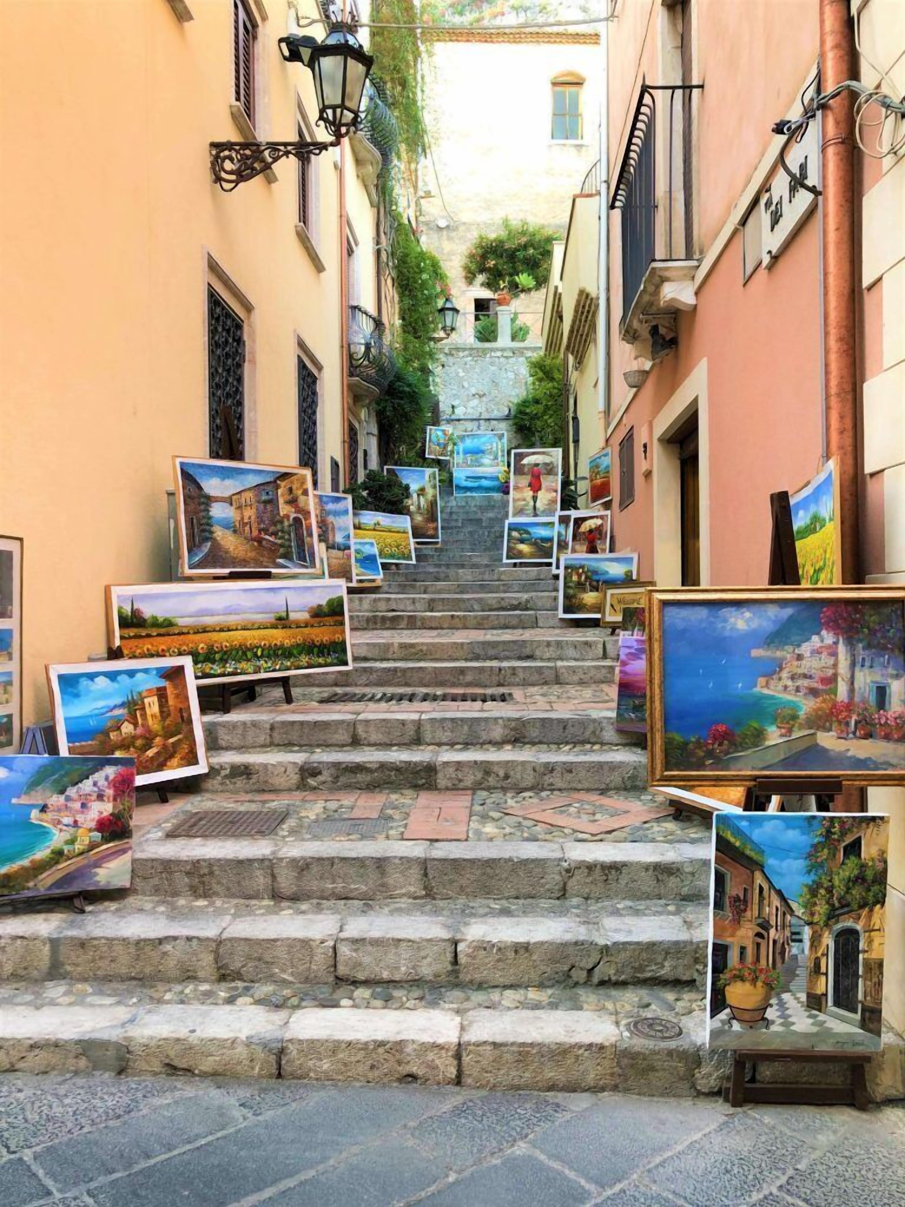 A narrow street with steps leading up to a building, adorned with numerous paintings.