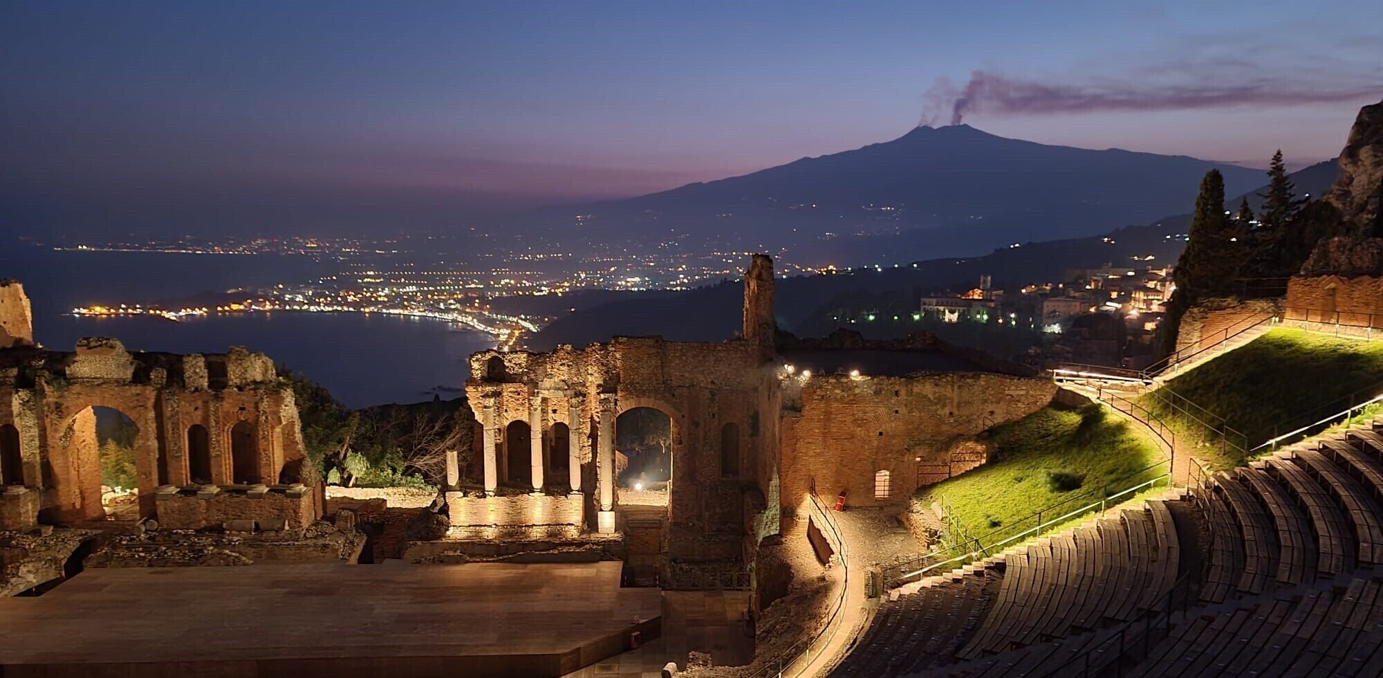 Un anfiteatro antiguo con ruinas y vistas a un paisaje urbano y un volcán al fondo.