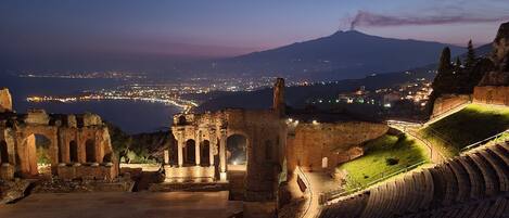 Un anfiteatro antiguo con ruinas y vistas a un paisaje urbano y un volcán al fondo.