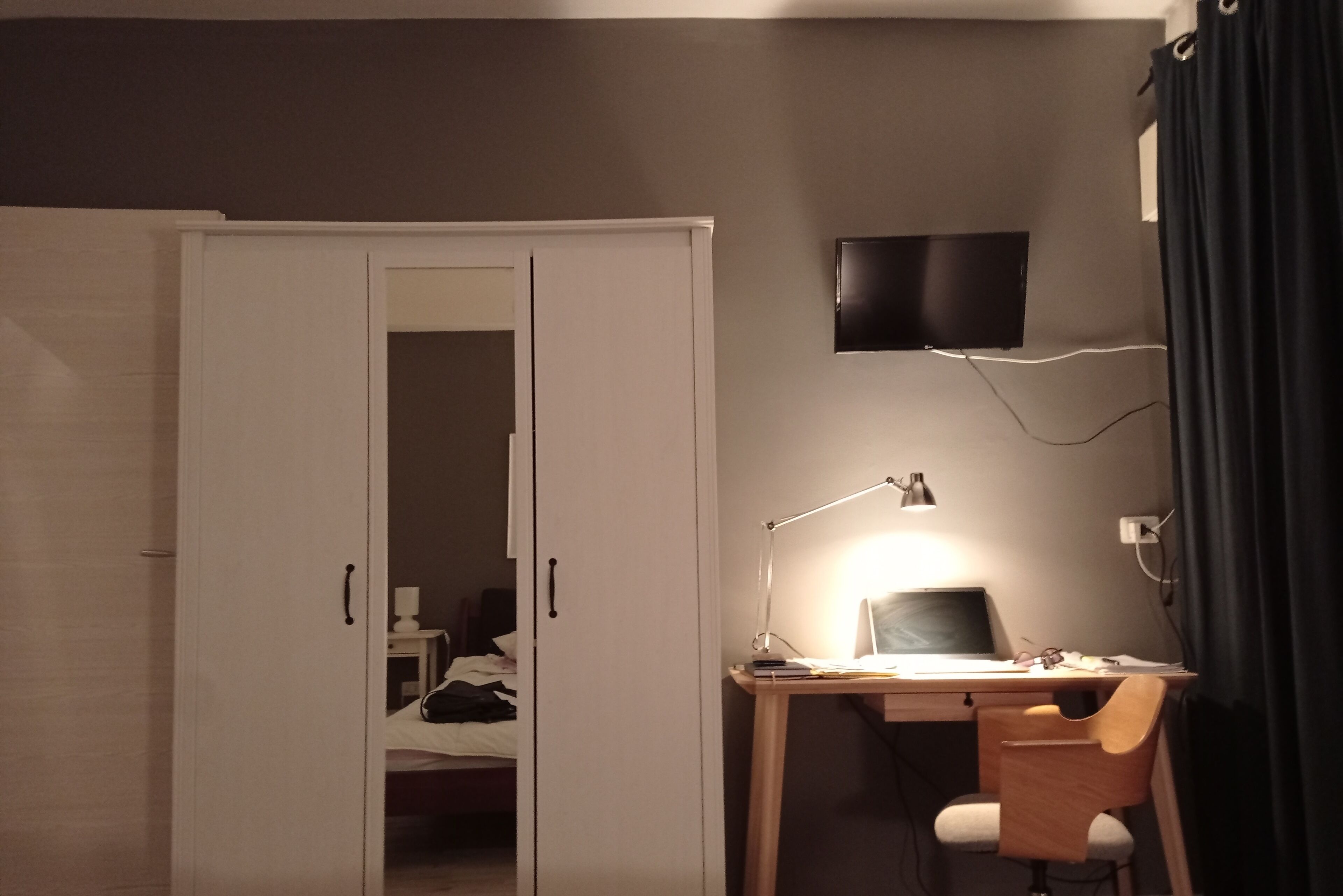 A bedroom with a bed, a desk with a computer, a chair, a desk lamp, and a TV on the wall.