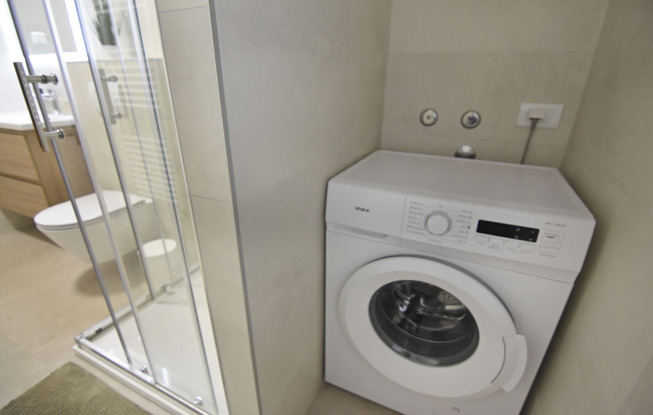 A compact bathroom with a glass shower enclosure, a white washing machine, and a toilet.