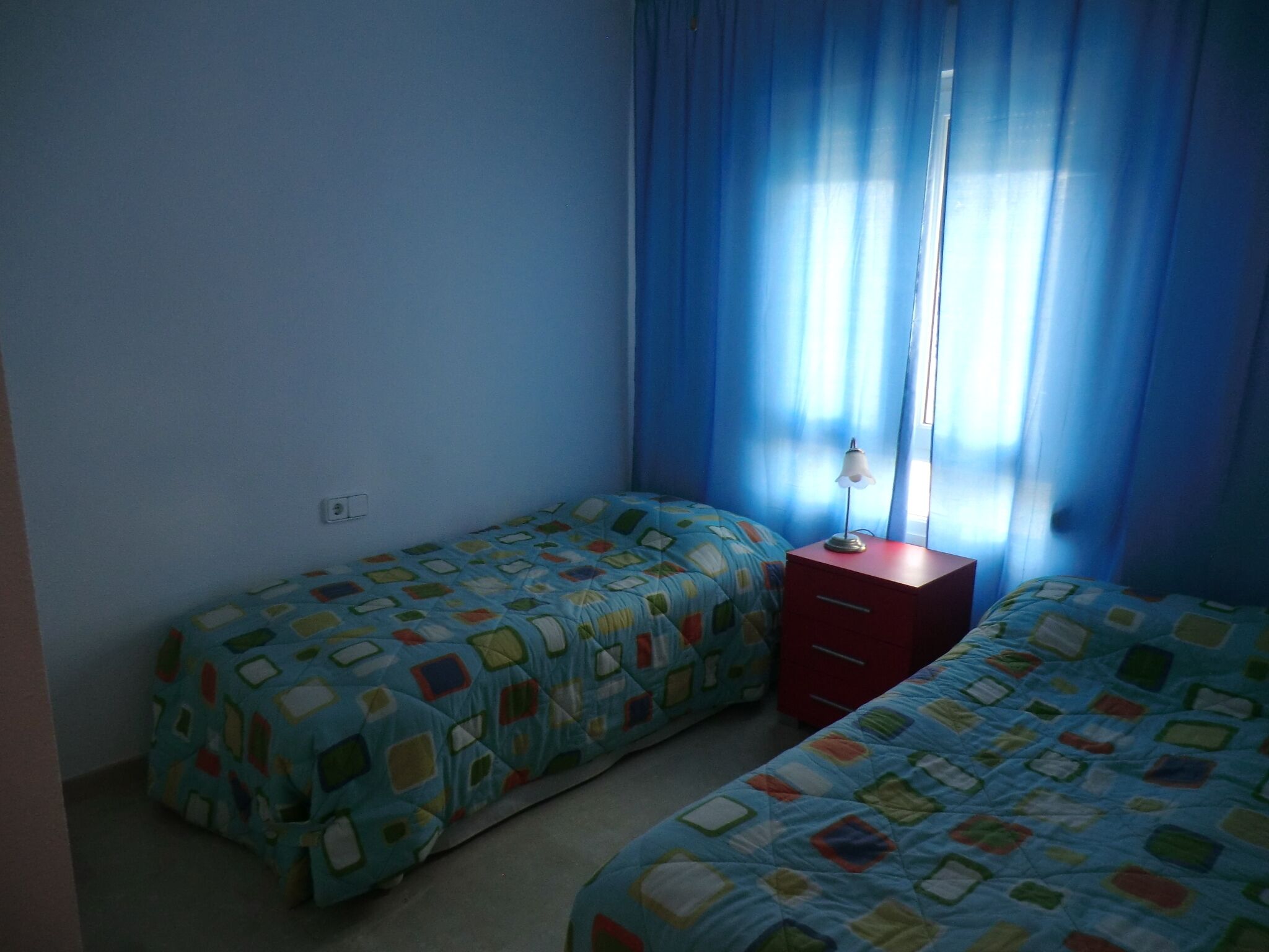 A room with two single beds, a red nightstand with a lamp, and a window with blue curtains.
