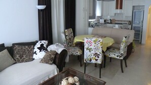Un salon comprenant un canapé, des fauteuils, une table basse et une table à manger recouverte de nappes à motifs floraux. Au fond, on aperçoit une cuisine avec des meubles blancs et des appareils électroménagers.