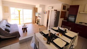 A modern living room with a sofa, dining table set for two, and a kitchen area with a refrigerator and oven. - Three bedroom apartment with sea views (Empuriabrava)