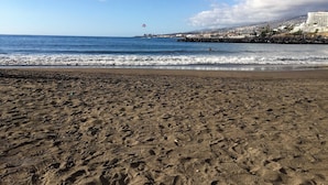 A beach with footprints leading to the water, a clear sky, and a distant view of a resort with buildings and a mountain. - Cozy and comfortable, best location! Ideal for families. (Costa Adeje)