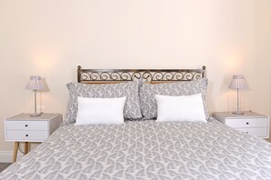 Een net opgemaakt bed met een bedsprei met patroon, twee witte kussens en een decoratief metalen hoofdbord. Er staan twee nachtkastjes met lampen en een klein ladeblok.