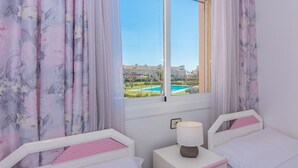 Ein Schlafzimmer mit zwei Betten, einem Fenster mit Blick auf einen Pool und Gebäude, sowie blumigen Vorhängen.