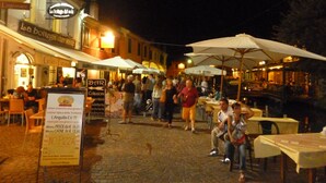 An outdoor dining area with tables and umbrellas, surrounded by buildings with signs indicating restaurants and cafes. - HOLIDAY RESIDENCE: sea and shopping within reach. (Lido degli Estensi)