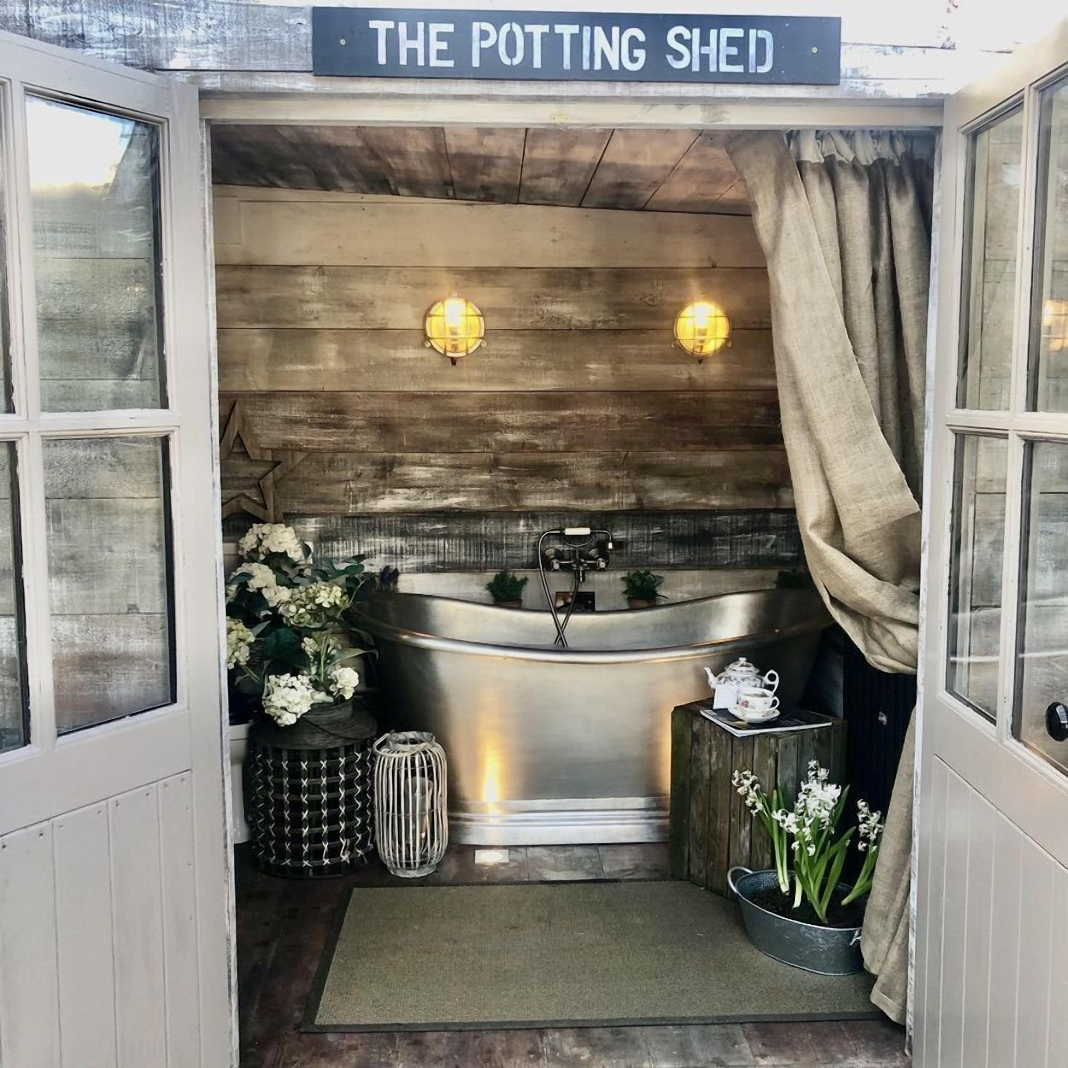 A rustic bathroom with a freestanding bathtub, wooden wall paneling, two wall-mounted lights, and a sign reading "THE POTTING SHEED".