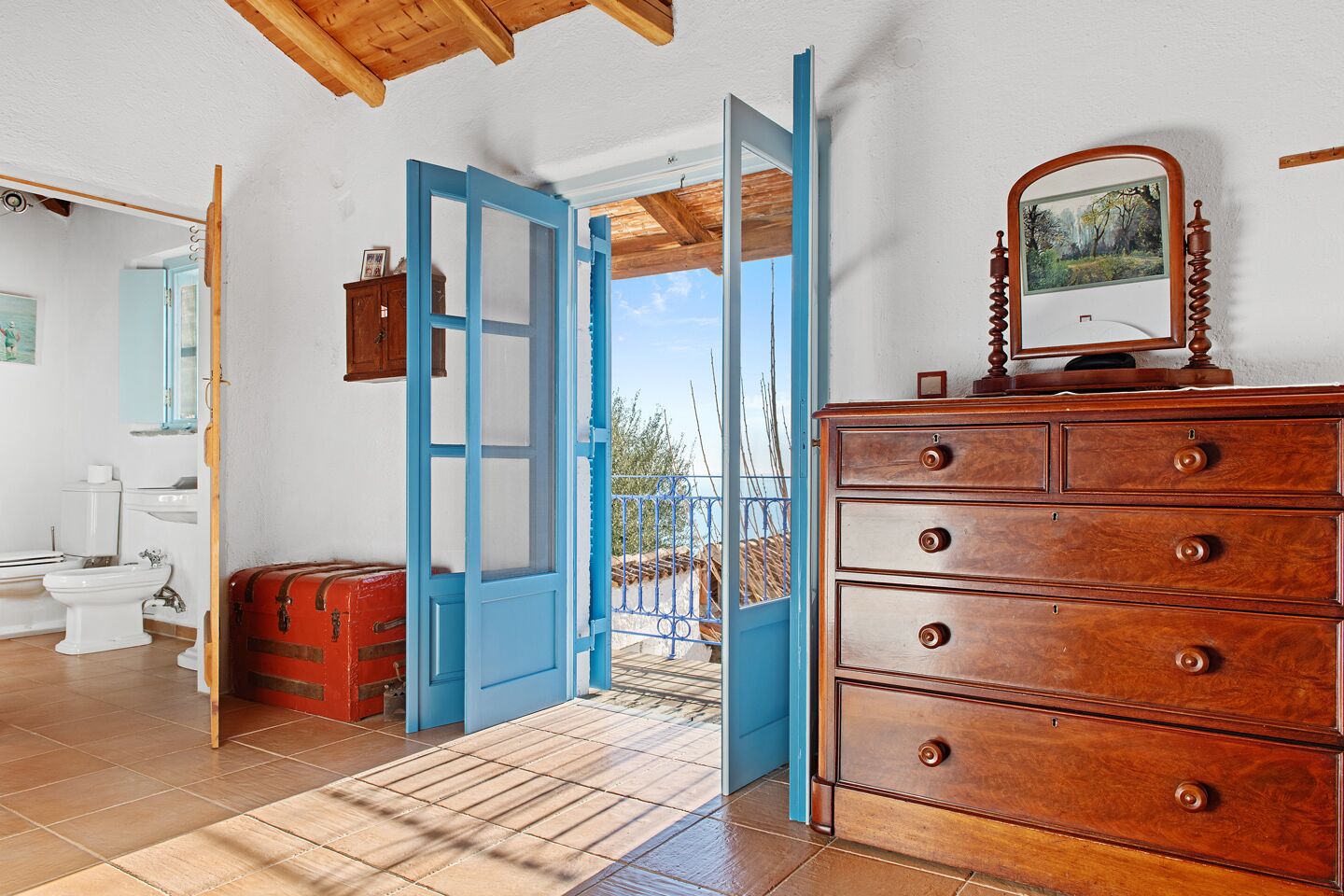 Une pièce avec une commode en bois, une valise rouge, une porte bleue donnant sur l’extérieur et une vue sur le paysage.