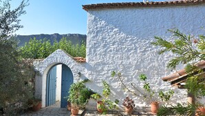 Un bâtiment blanc avec une porte bleue, entouré de plantes en pot et d’une cour dallée de pierres.