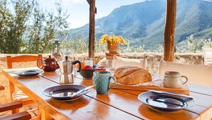 Une table en bois, dressée avec des assiettes, une théière, un pichet, une tasse et un pain, d’où l’on peut admirer un paysage montagneux.