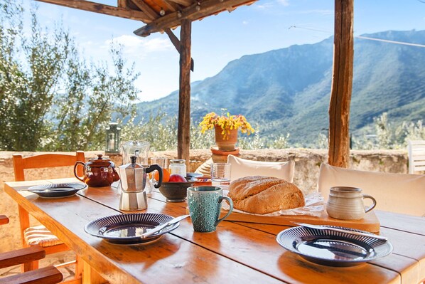 A wooden table set with plates, a teapot, a pitcher, a mug, and a loaf of bread, overlooking a mountainous landscape.