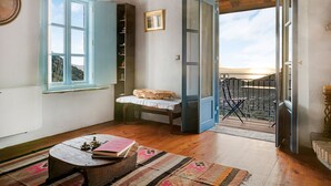 Une pièce chaleureuse avec un plancher en bois, un lit recouvert d’une courtepointe blanche, une petite table avec des livres, une fenêtre avec des volets bleus et un balcon doté d’une rambarde en métal.