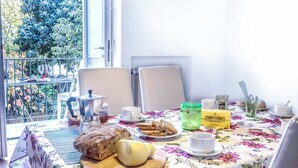 Una mesa de comedor con mantel floral, hogaza de pan, queso redondo y diversos elementos para el desayuno, incluyendo una cafetera, frascos y una caja de té Earl Grey.