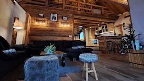 Un interior rústico con vigas de madera a la vista, una zona de estar acogedora con sofá y reposapiés, un comedor con barra y una cocina al fondo.