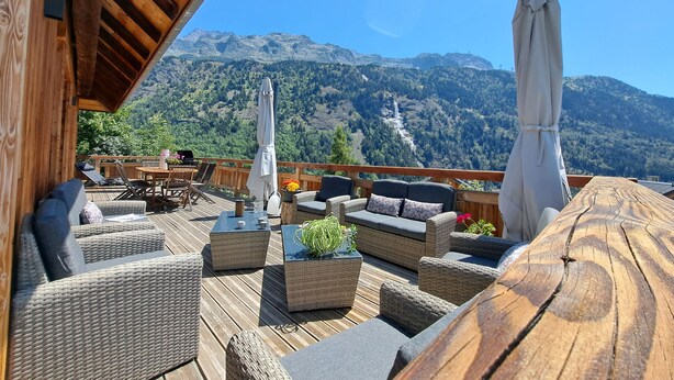 Una terraza de madera con muebles de mimbre y sombrillas, con vista a un paisaje montañoso.