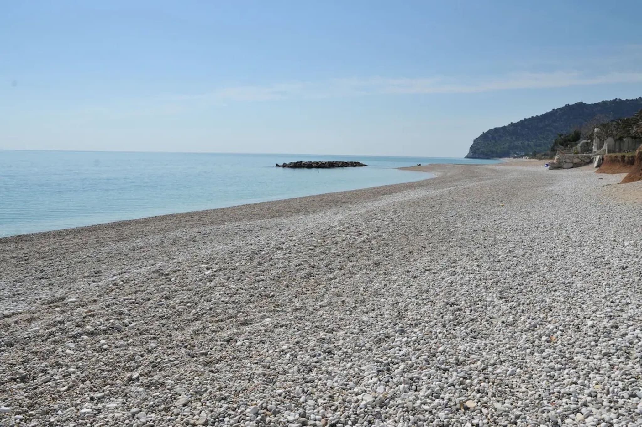 Platja de còdol amb un promontori rocosos a l'aigua, zona sorrenca amb un turó al fons i cel serè.