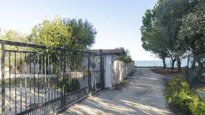 Un sendero pavimentado conduce a una entrada con puerta, una pared blanca y una estructura con techo de tejas. Árboles bordean el camino, y al fondo se ve un cielo despejado y una masa de agua.
