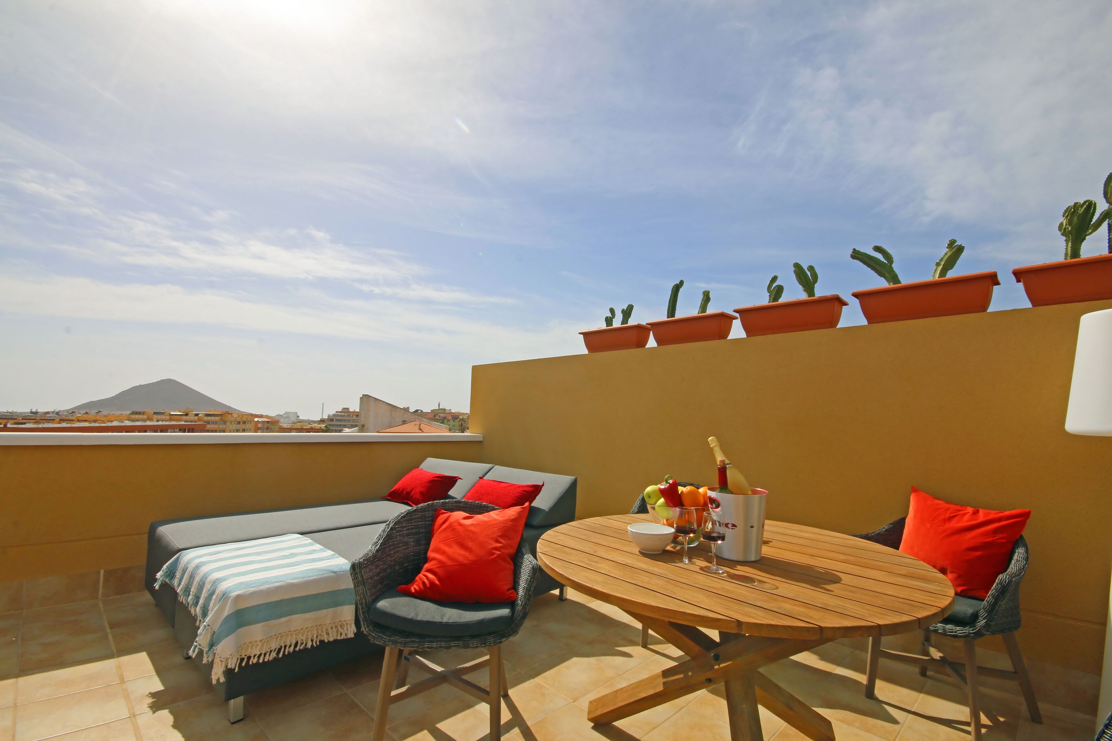 A rooftop terrace with a round wooden table, two wicker chairs, a sofa with red cushions, and potted cacti.