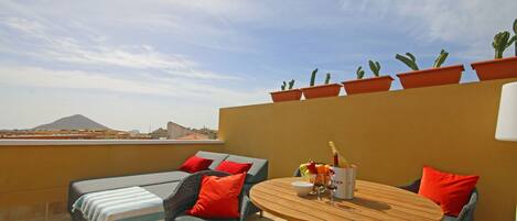A rooftop terrace with a round wooden table, two wicker chairs, a sofa with red cushions, and potted cacti.