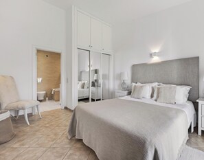 Une chambre à coucher avec un lit, des tables de chevet, une chaise, une armoire et une salle de bain visible à travers une porte ouverte.