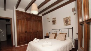 Un dormitorio con un armario de madera grande, una cama con cubrecama blanco, dos lámparas de pared y un ventanal con marcos de madera.