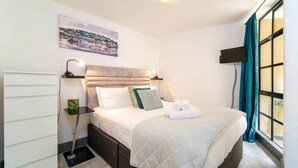 A neatly arranged hotel room with a bed, a bedside table with a lamp, a flat-screen TV, and a window with curtains. - Elegant apartment near St Paul’s (London)