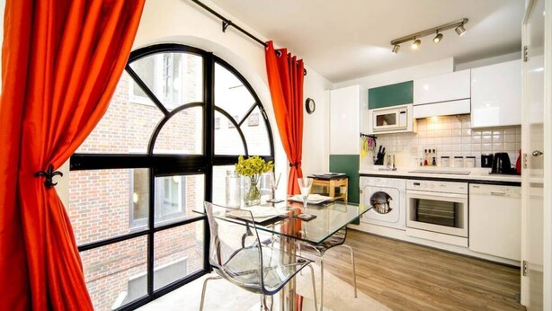A modern kitchen with white cabinets, a microwave, and a sink. There is a dining table with chairs and a window with curtains. - Elegant apartment near St Paul’s (London)