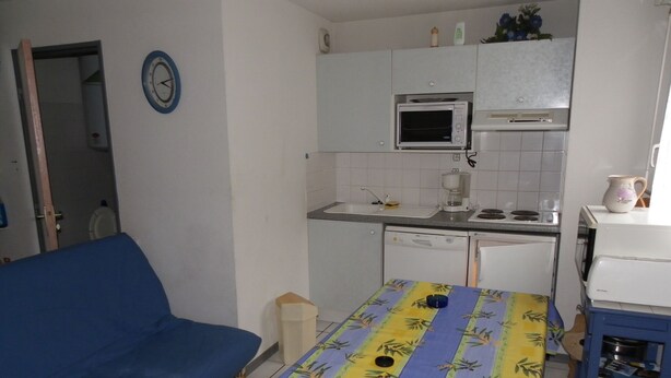 Cocina compacta con microondas, estufa y lavavajillas. Hay un sofá azul y una mesa con mantel floreado.