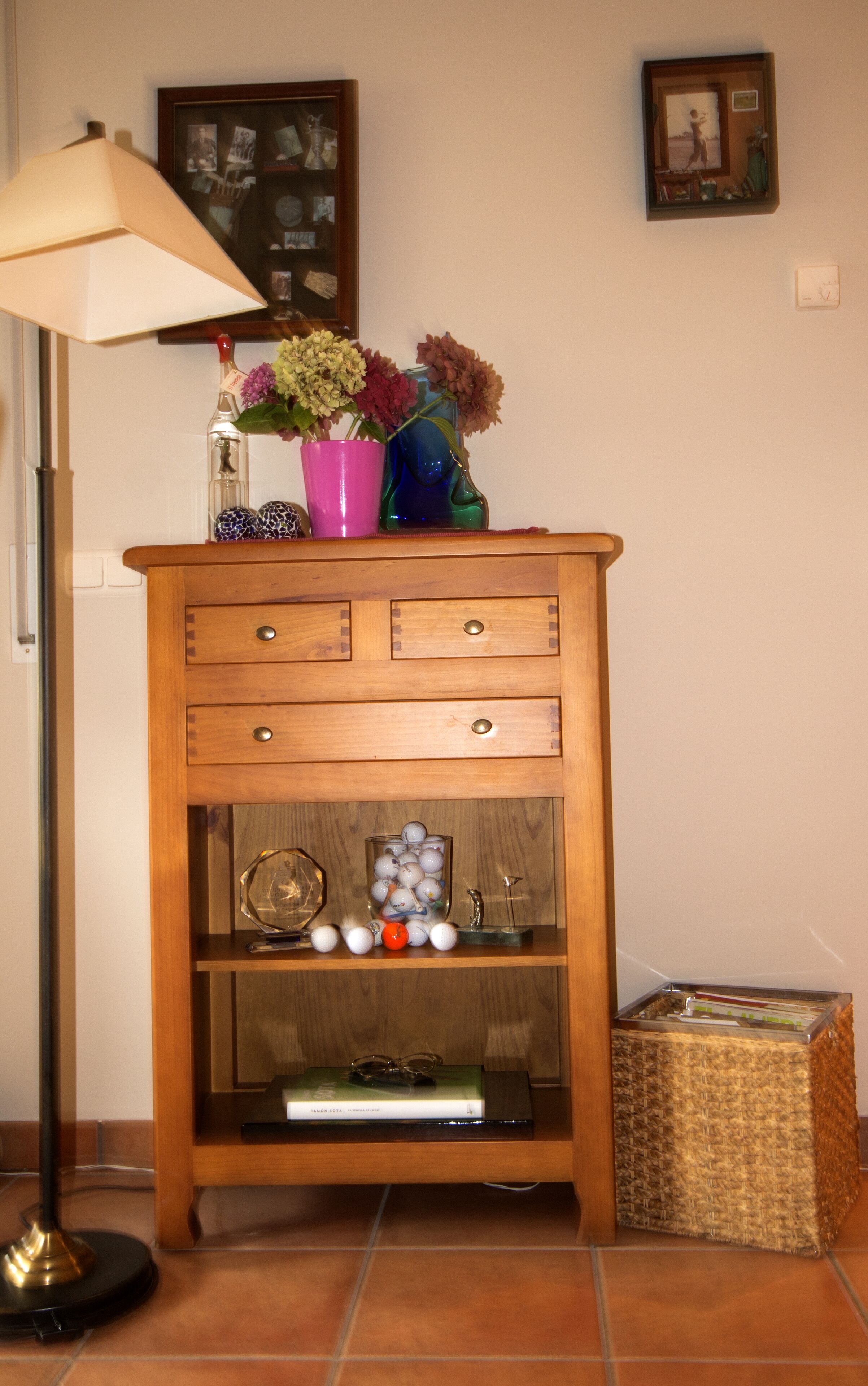A wooden cabinet with three drawers, a shelf with decorative items, and a lamp to the left.