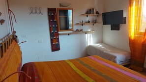 A room with a bed, a TV mounted on the wall, a shelf with books and decorative items, and a window with curtains.