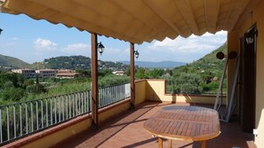 A balcony with a table and chairs, a railing, and a view of a hilly landscape with buildings in the distance.