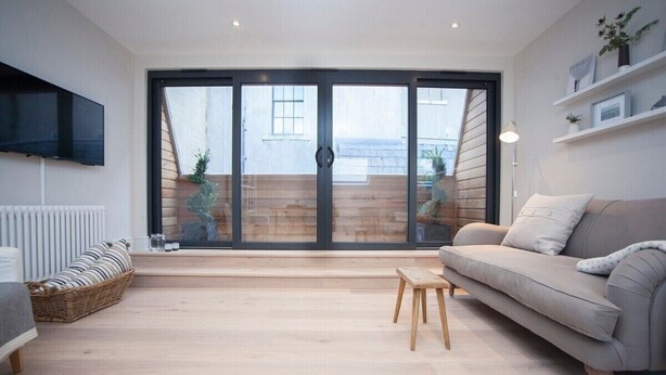 A modern living room with a sofa, a small wooden stool, a television mounted on the wall, and a sliding glass door leading to an outdoor area. - Stylish Apt in Heart of Georgian Bath (WST) (Bath)