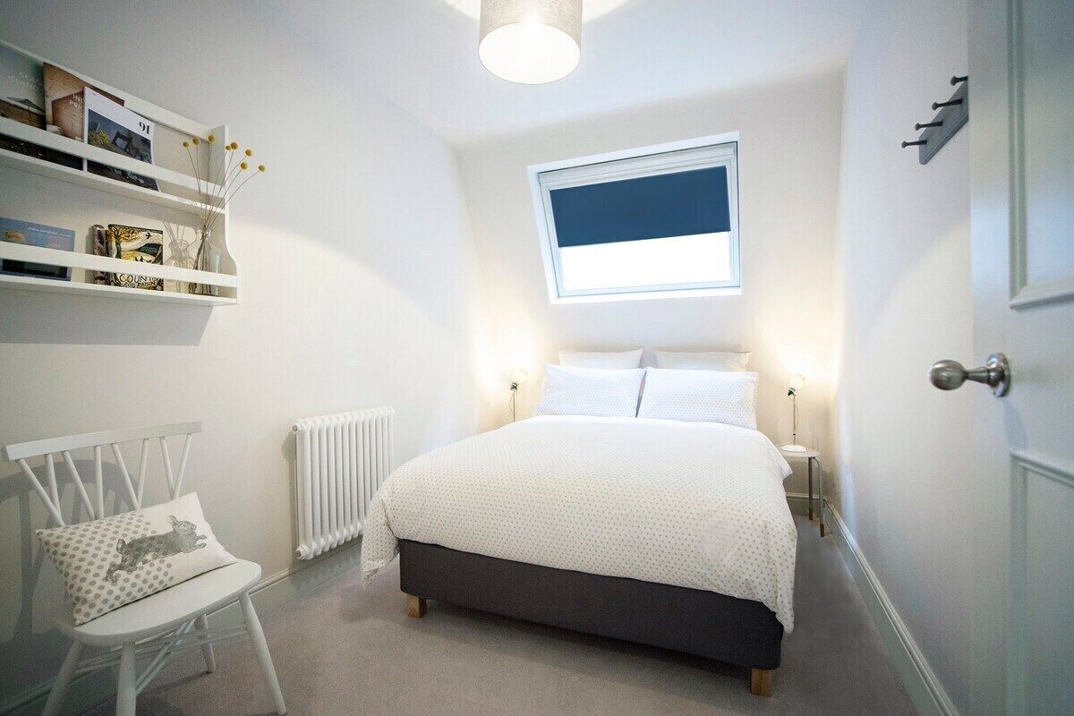 A bedroom with a bed, a chair, a radiator, a shelf with books, and a window.