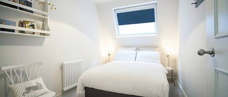A bedroom with a bed, a chair, a radiator, a shelf with books, and a window.